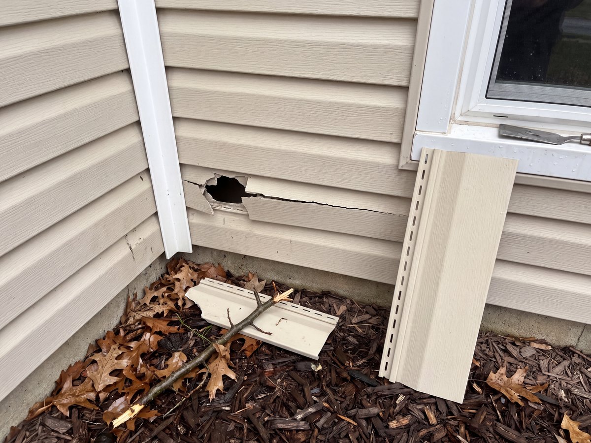 Beige vinyl siding with a storm-cracked panel near an inside corner, the broken piece and a replacement panel beside it