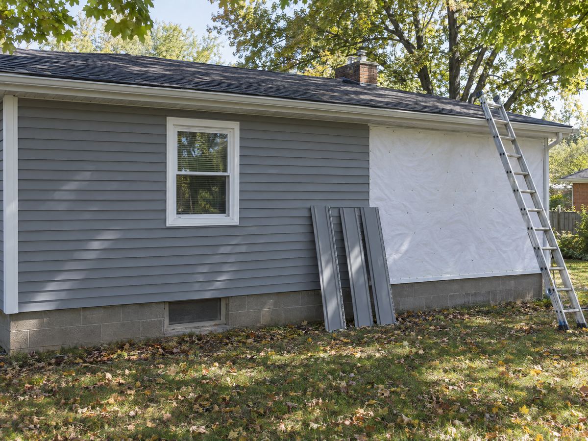 Ranch house wall two-thirds sided in new gray vinyl, the rest in house wrap, with panels and a ladder leaning on it